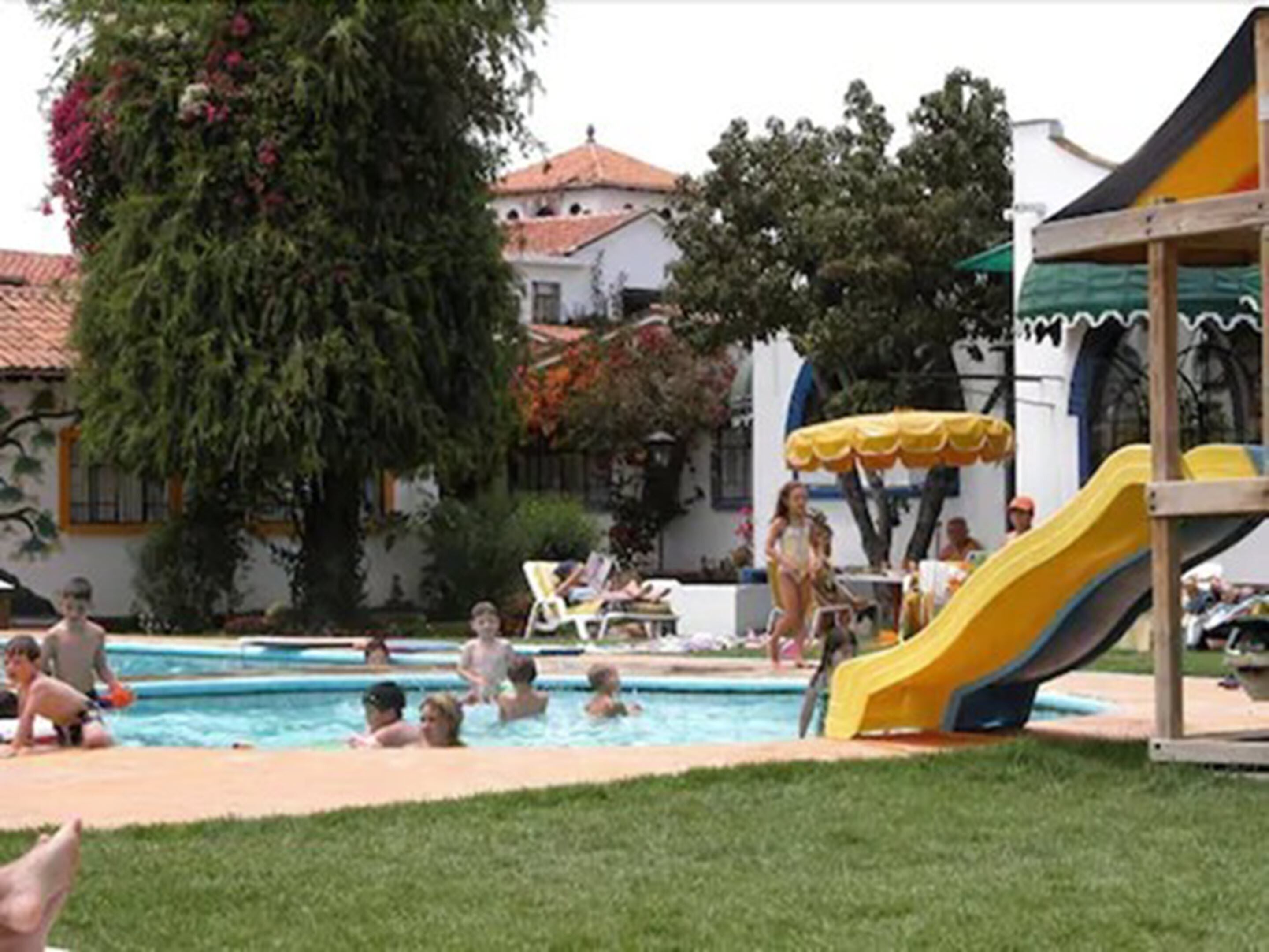 Piscina com escorrega, cercada por vegetação e um edifício com telhado de azulejos.