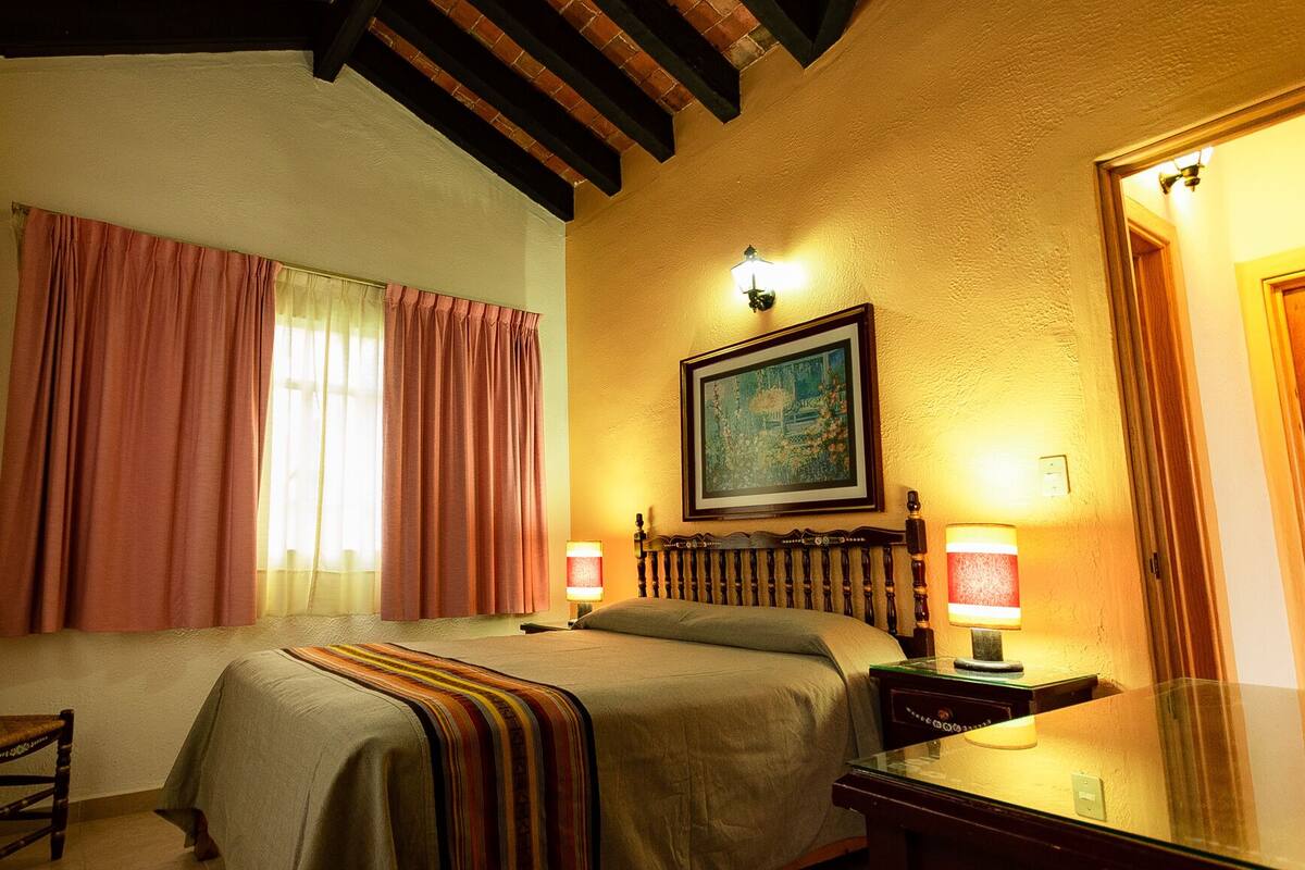 A hotel room with a bed, two bedside lamps, a window with curtains, a painting on the wall, and a mirror.
