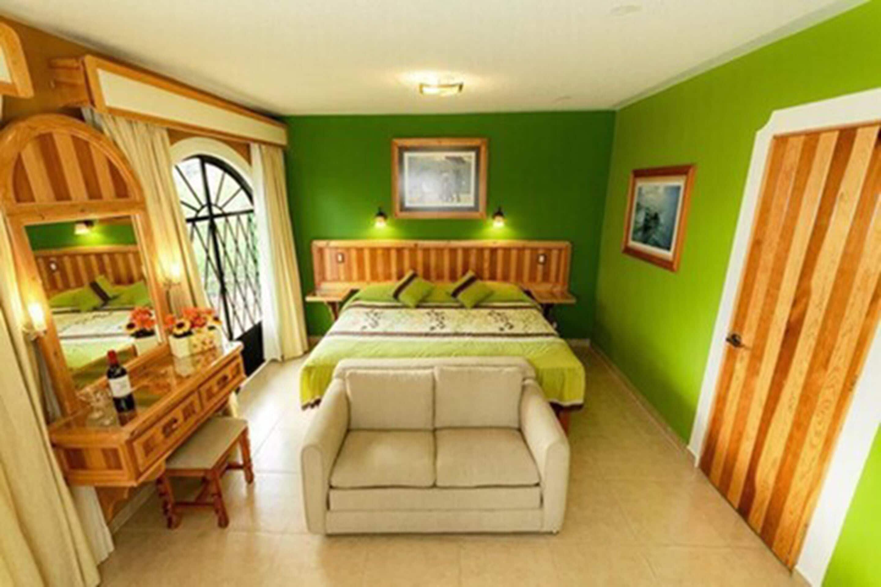 Une chambre à coucher avec un lit, une commode en bois, un miroir, un banc et un mur vert.