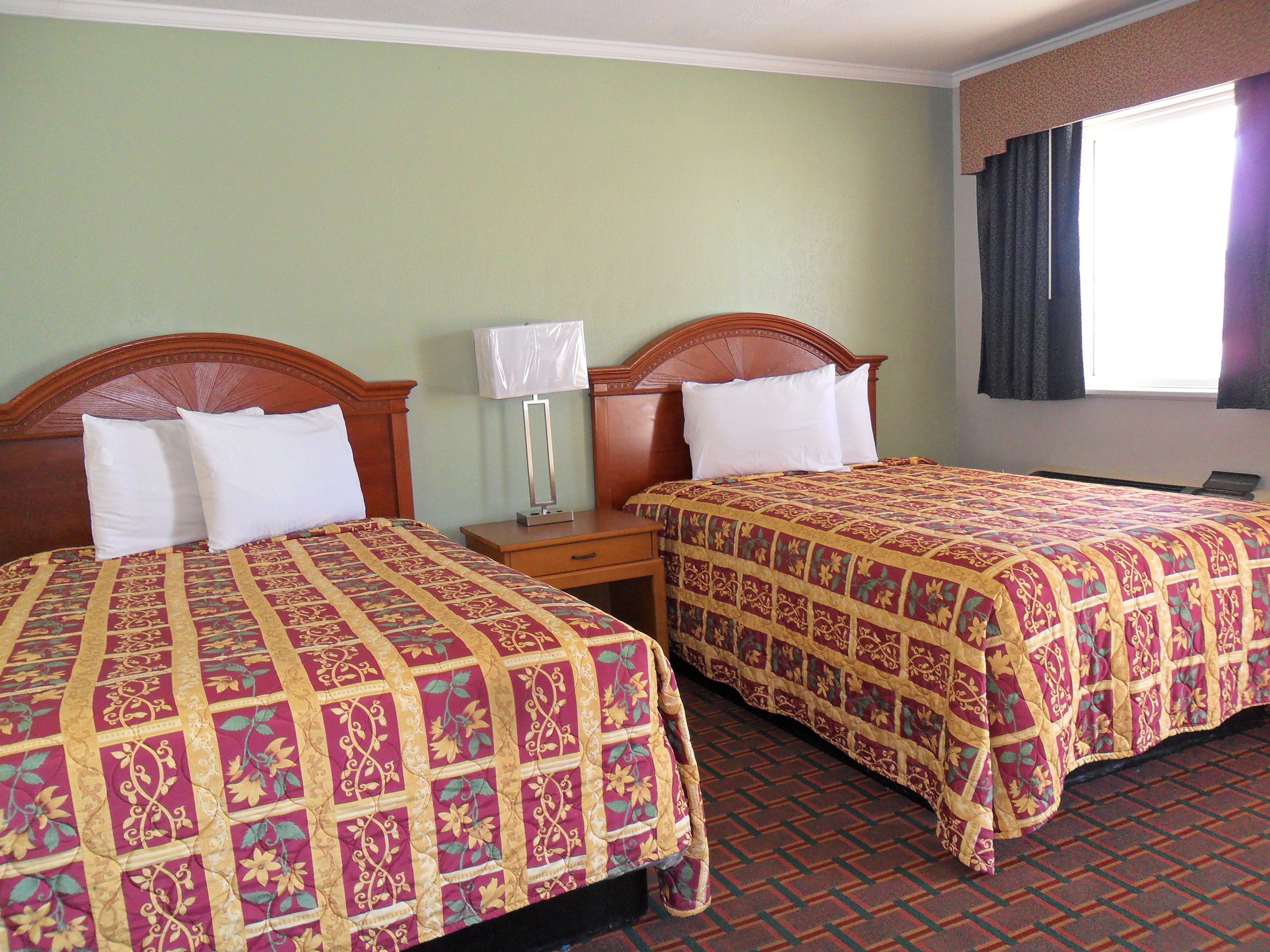 A hotel room with two beds, each with a floral-patterned bedspread, a bedside table with a lamp, and a window with curtains.