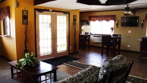 A living room with a sofa, a dining table, and a TV. There are two windows with blinds, a chandelier, and a plant. - Country Inn Sterling (Sterling)
