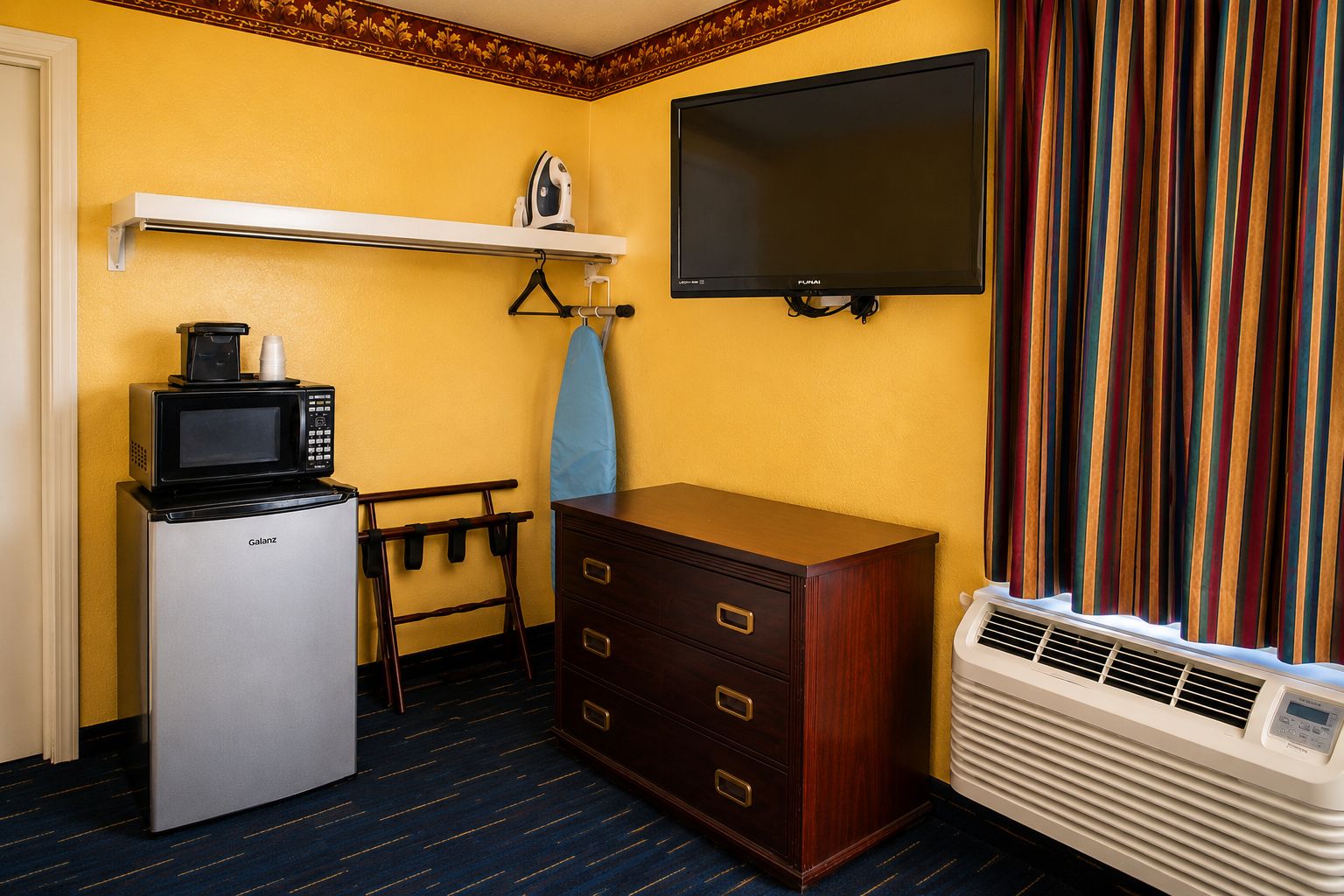 Ein kompaktes Hotelzimmer mit Küchenzeile, einem Flachbildfernseher, einem Holzschrank, einer Bank und einem Fenster mit Vorhängen.