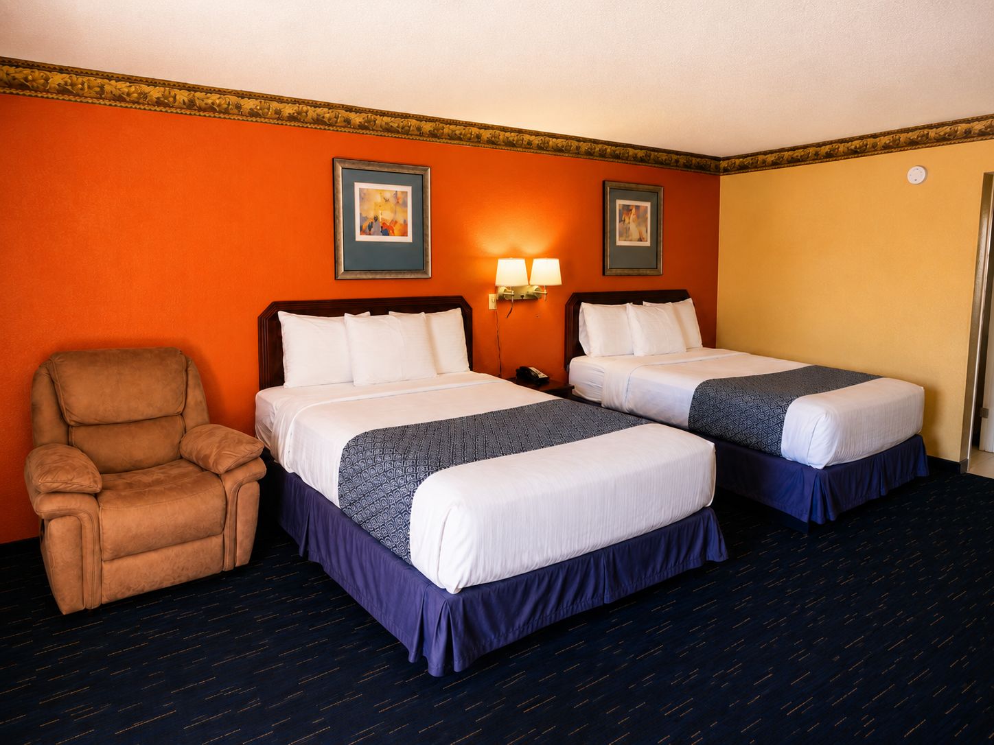 Quarto de hotel com duas camas, uma cadeira e duas luminárias fixadas na parede.