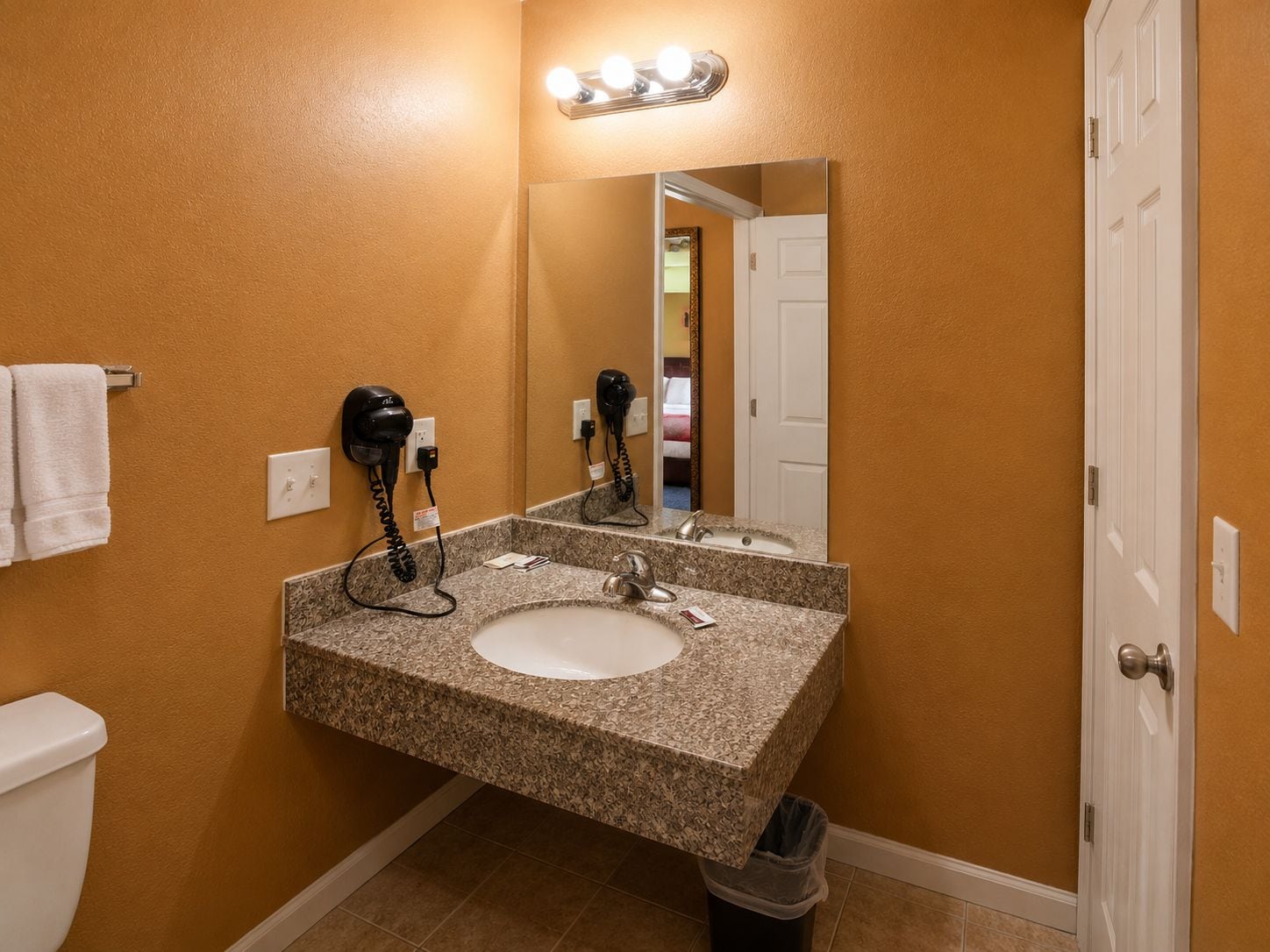 A bathroom with a granite sink, a hairdryer, a mirror, and a trash can.