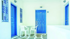Un pasillo estrecho con una puerta azul identificada con el número 21, paredes blancas y una pequeña mesa con dos sillas de plástico blancas.