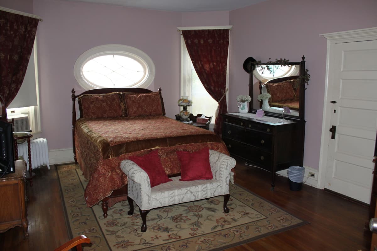 A bedroom with a large bed, a dresser, a vanity with a mirror, and a small white chair.