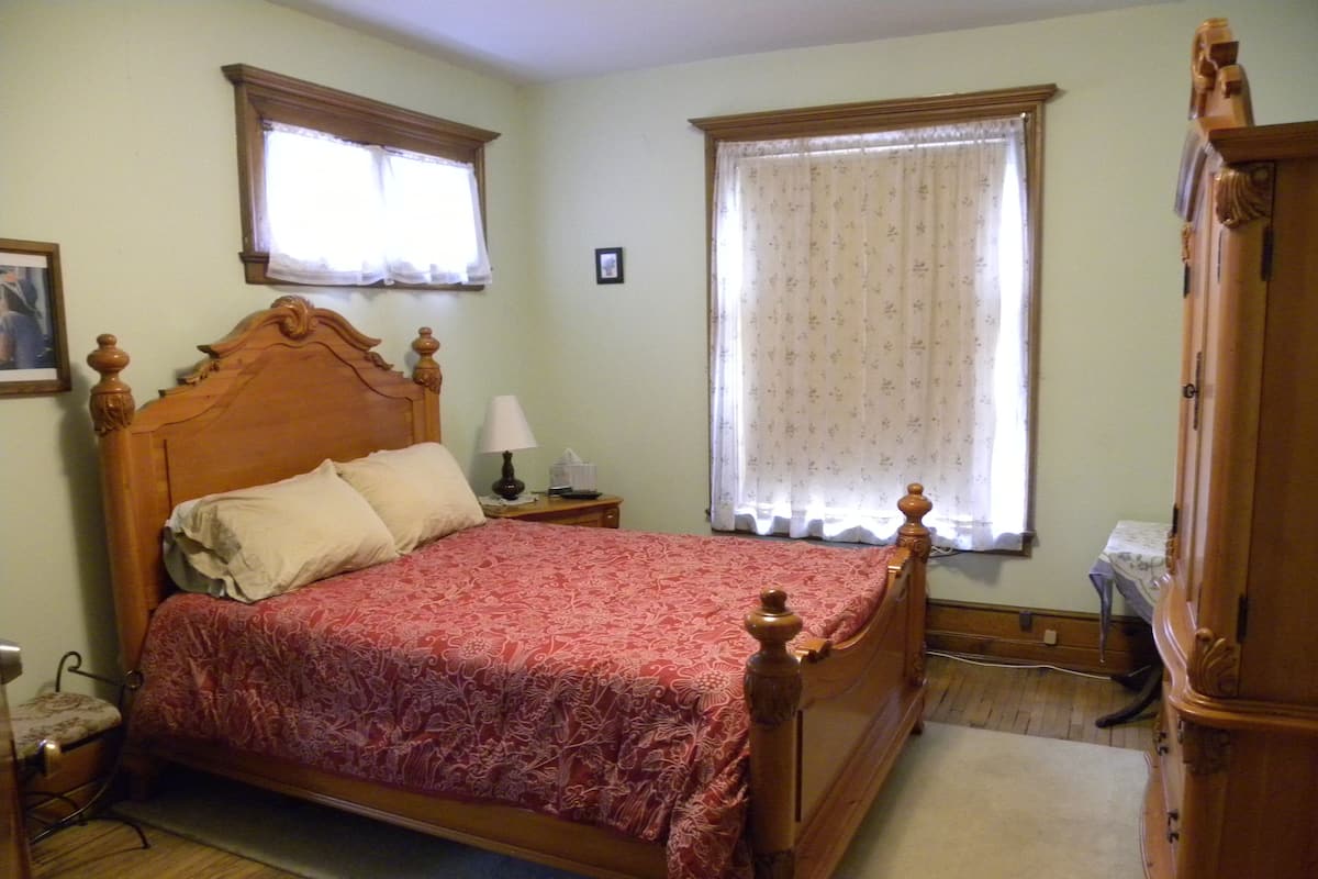 A bedroom with a wooden bed, two windows with curtains, a nightstand, and a dresser.