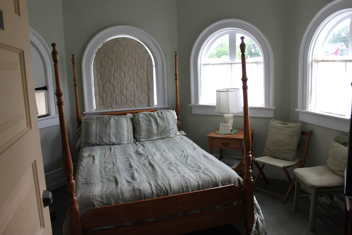 A four-poster bed with a wooden frame and a grey striped bedspread. There are two windows with white frames and a view of the outside. A bedside table with a lamp and a chair is visible.