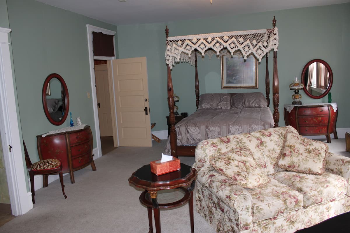 A bedroom with a four-poster bed, a floral sofa, a wooden dresser, a mirror, and a small table with a tissue box.
