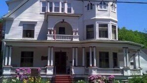 A two-story house with a central entrance, a porch with columns, and a small front yard with flowers.
