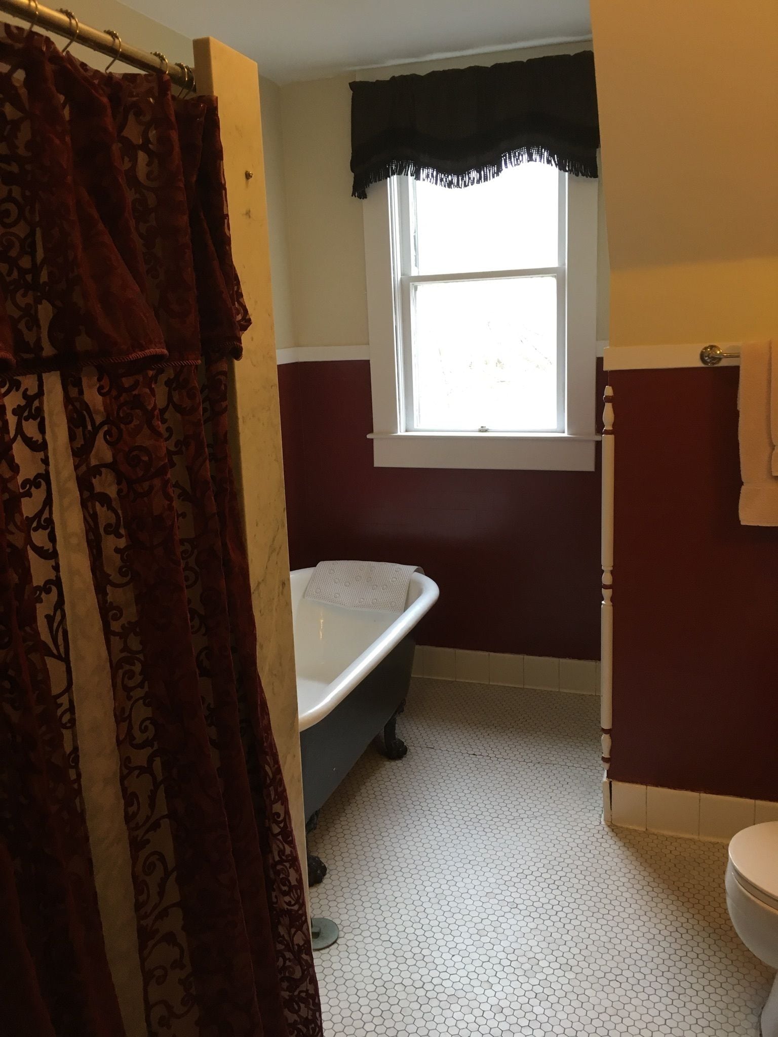 A bathroom with a clawfoot bathtub, a window with a black valance, and a white toilet.