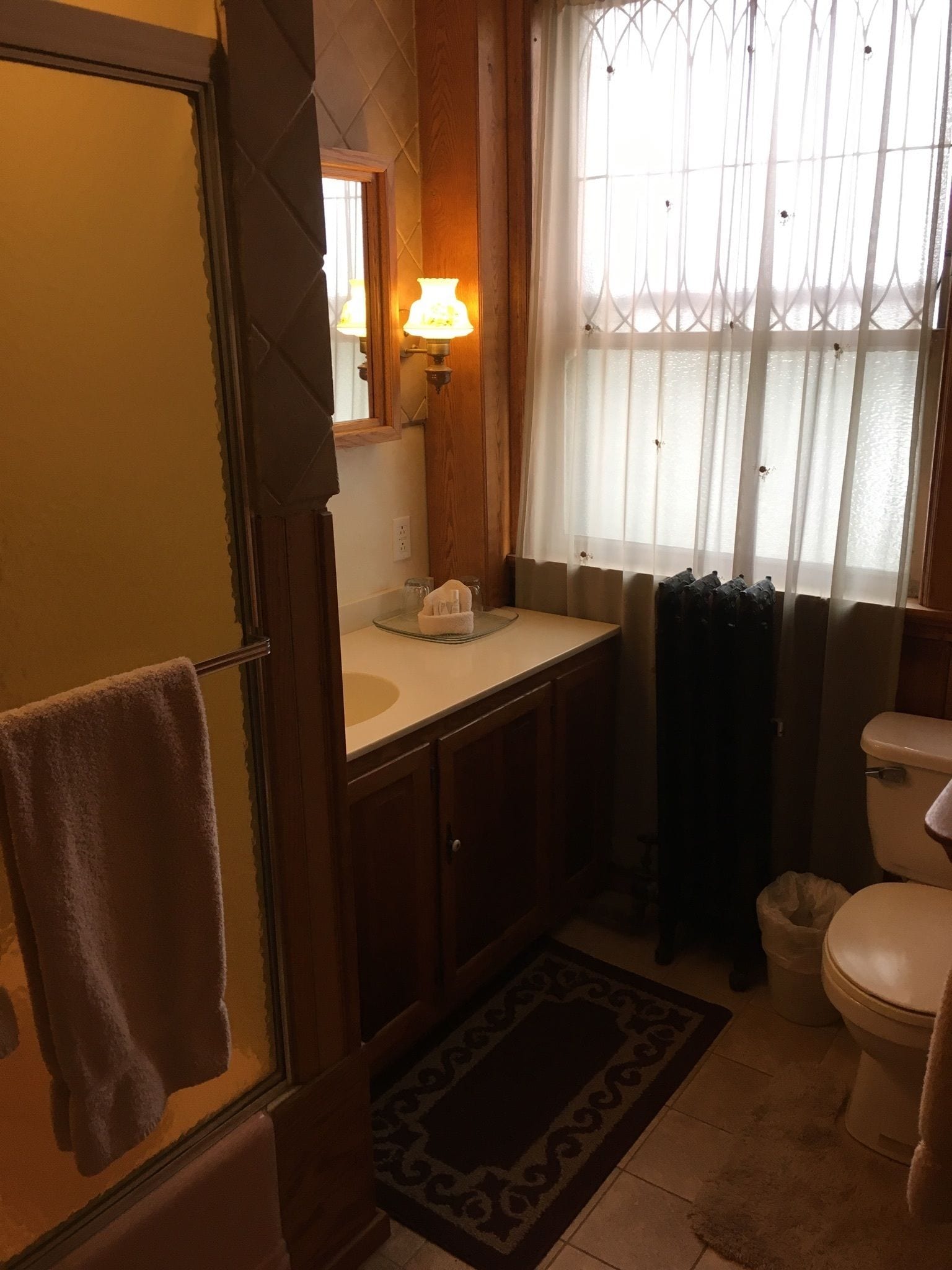 Un baño con mampara de vidrio, lavamanos con espejo, inodoro y ventana con cortina.