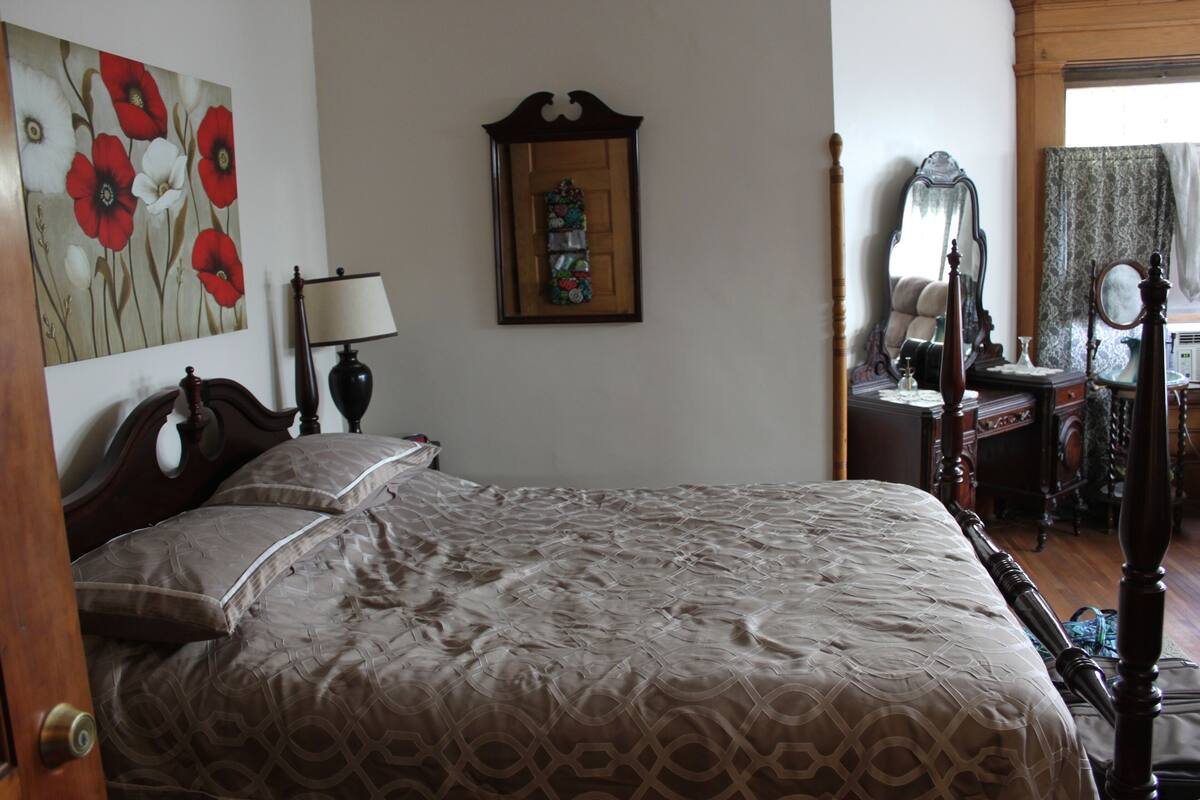 A bedroom with a large bed, a wooden headboard, a bedside table with a lamp, a dresser, a mirror, and a framed artwork on the wall.