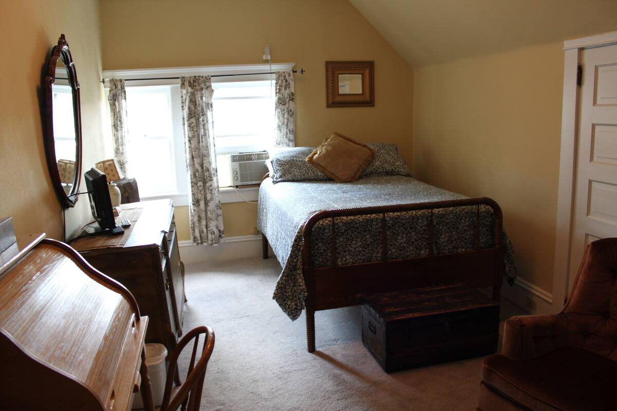 A bedroom with a bed, a wooden desk, a chair, a dresser, and a window with curtains.