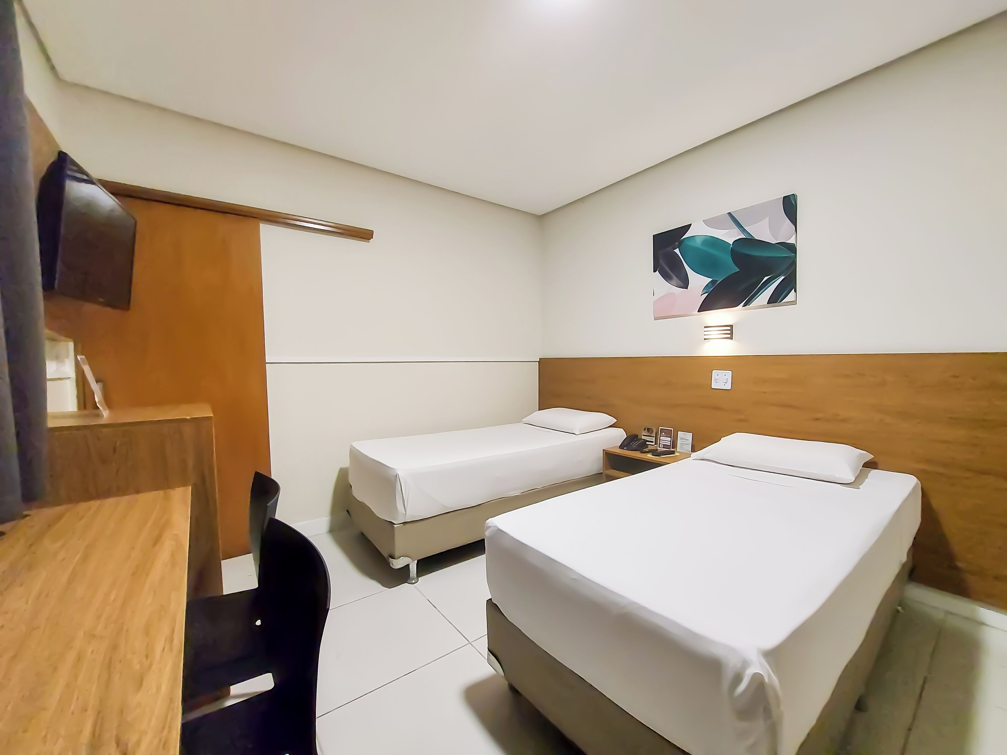 A hotel room with two single beds, a wooden desk, a chair, a bedside table with a phone and a lamp, and a wall-mounted artwork.