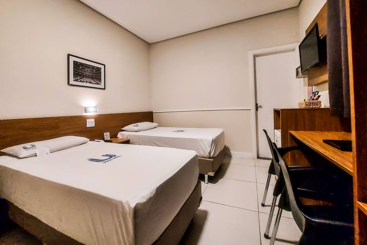 A hotel room with two single beds, a wooden desk, a chair, a television mounted on the wall, and a framed picture on the wall.
