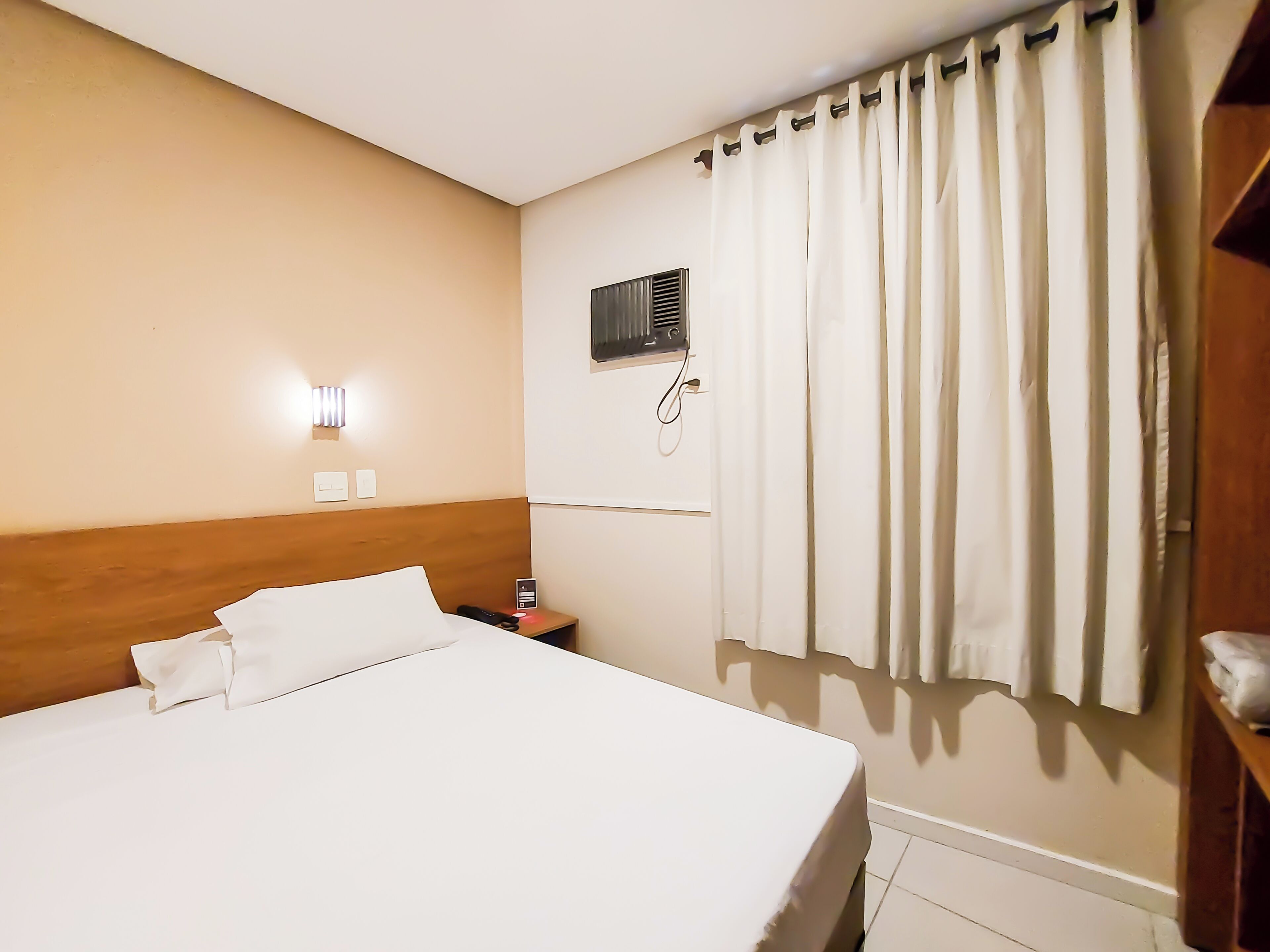 Ein Hotelzimmer mit einem ordentlich bezogenen Bett, einem Nachttisch mit Telefon, einer Wand-Klimaanlage und einem Fenster mit Vorhängen.