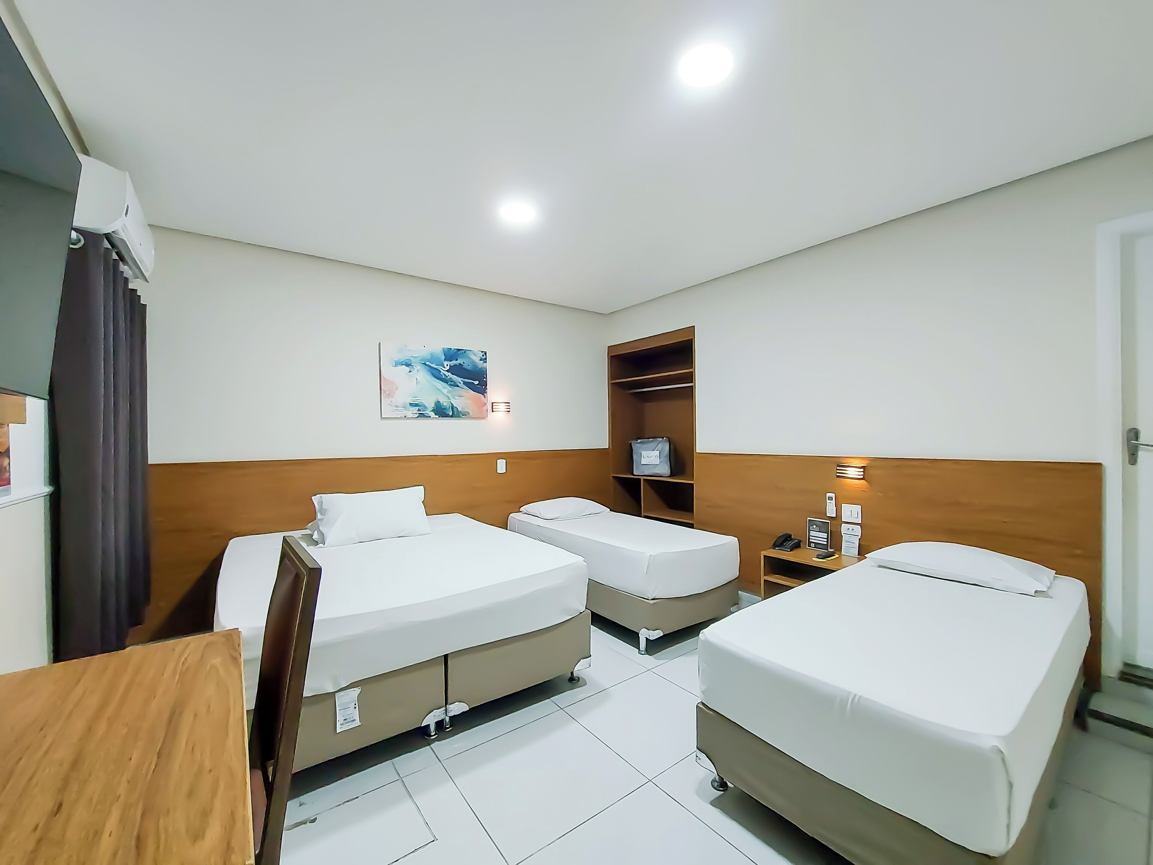 A hotel room with two single beds, a wooden headboard, a small desk, a nightstand, a wall-mounted TV, and a shelf with a bag.
