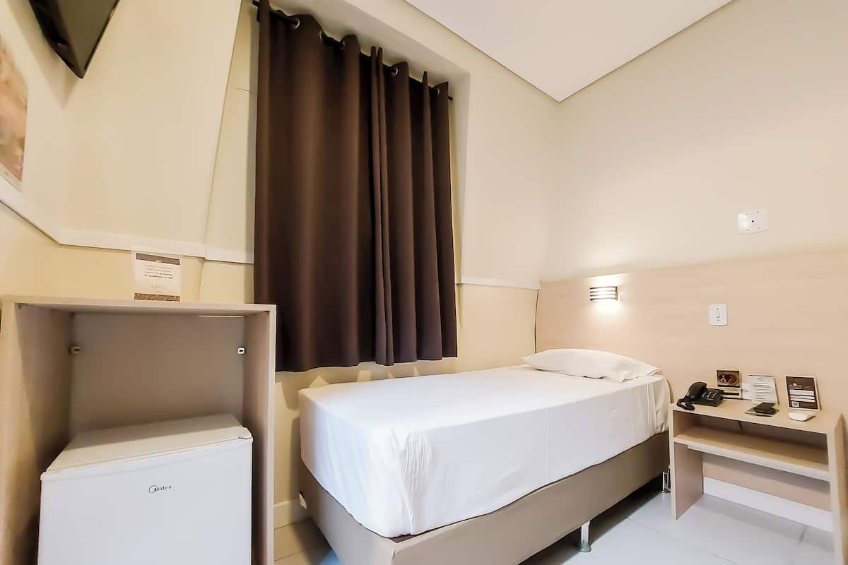 A hotel room with a bed, a small desk, a refrigerator, and a window with curtains.