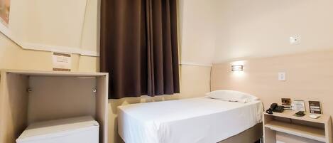A hotel room with a bed, a small desk, a refrigerator, and a window with curtains.