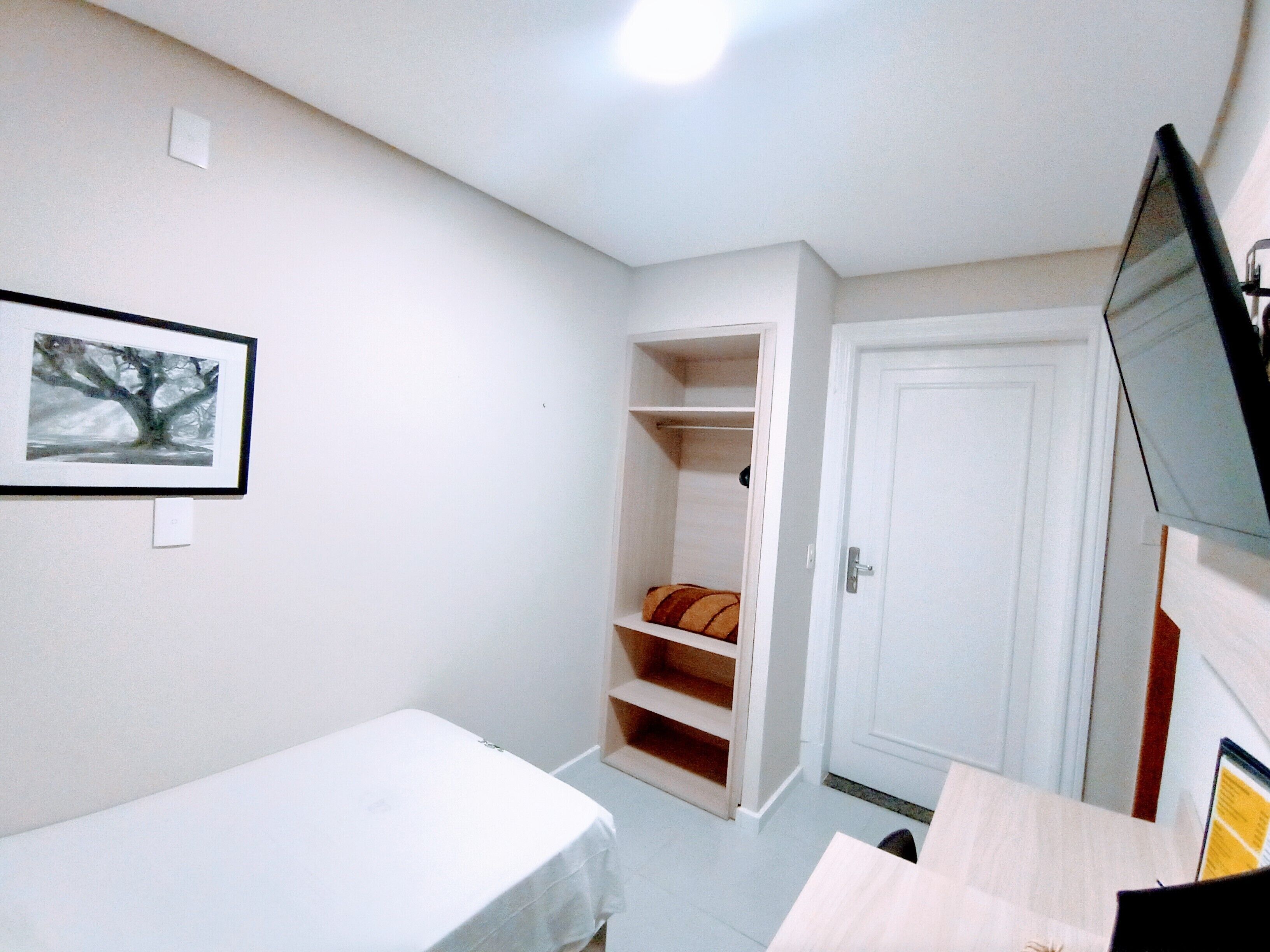 Una habitación pequeña y escasamente amoblada, con una cama, una puerta blanca, un estante con un cojín y un cuadro en la pared.