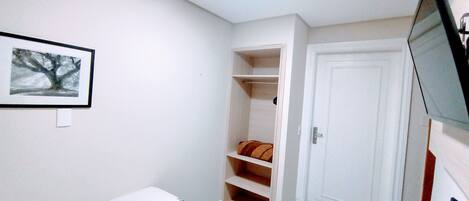 Una habitación pequeña y escasamente amoblada, con una cama, una puerta blanca, un estante con un cojín y un cuadro en la pared.