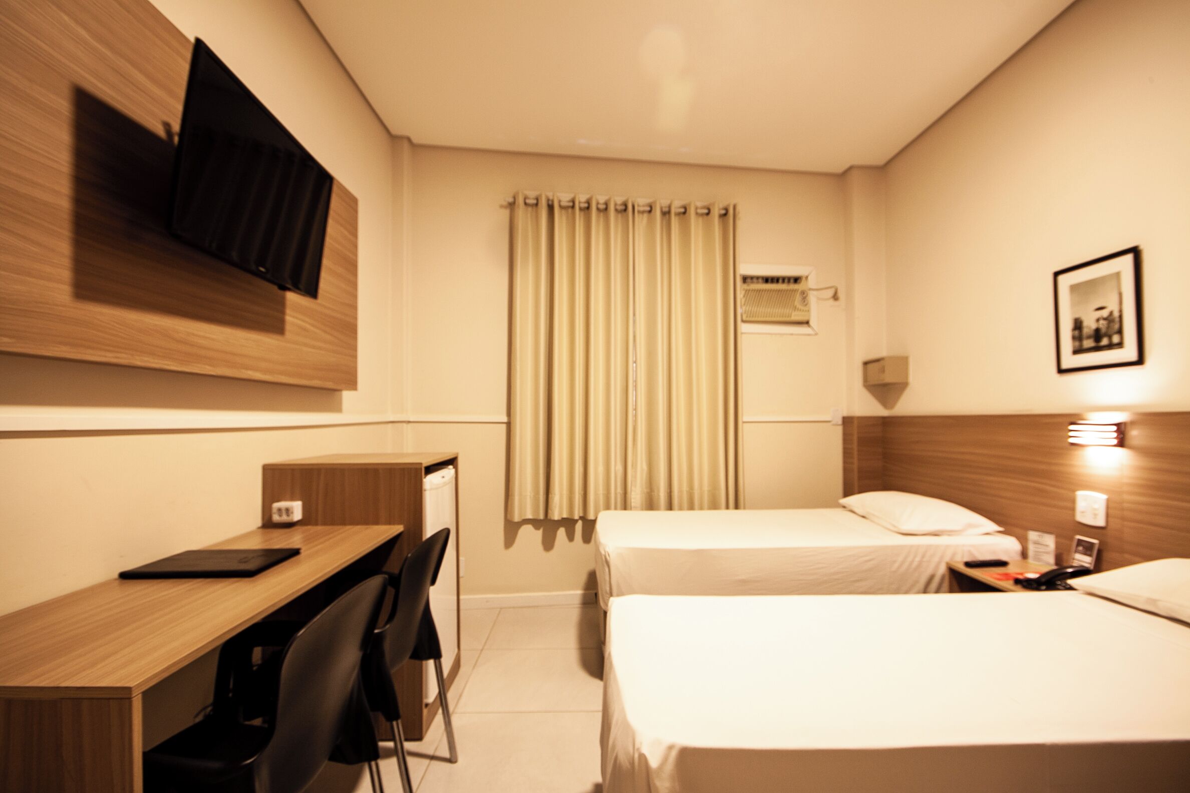 Quarto de hotel com uma escrivaninha de madeira, duas camas, uma televisão na parede, uma janela com cortinas e uma mesa de cabeceira com luminária.