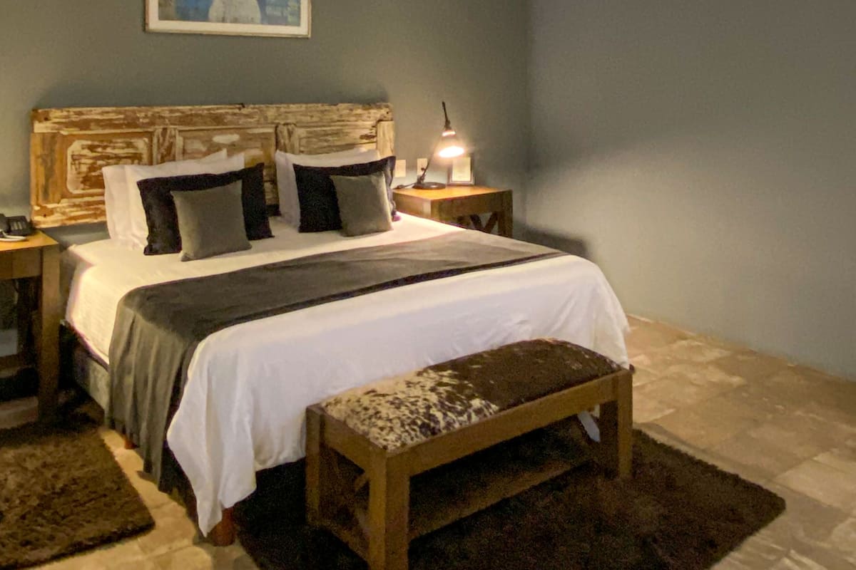 A bedroom with a large bed, a wooden headboard, a bedside table with a lamp, a bench, and a painting on the wall.