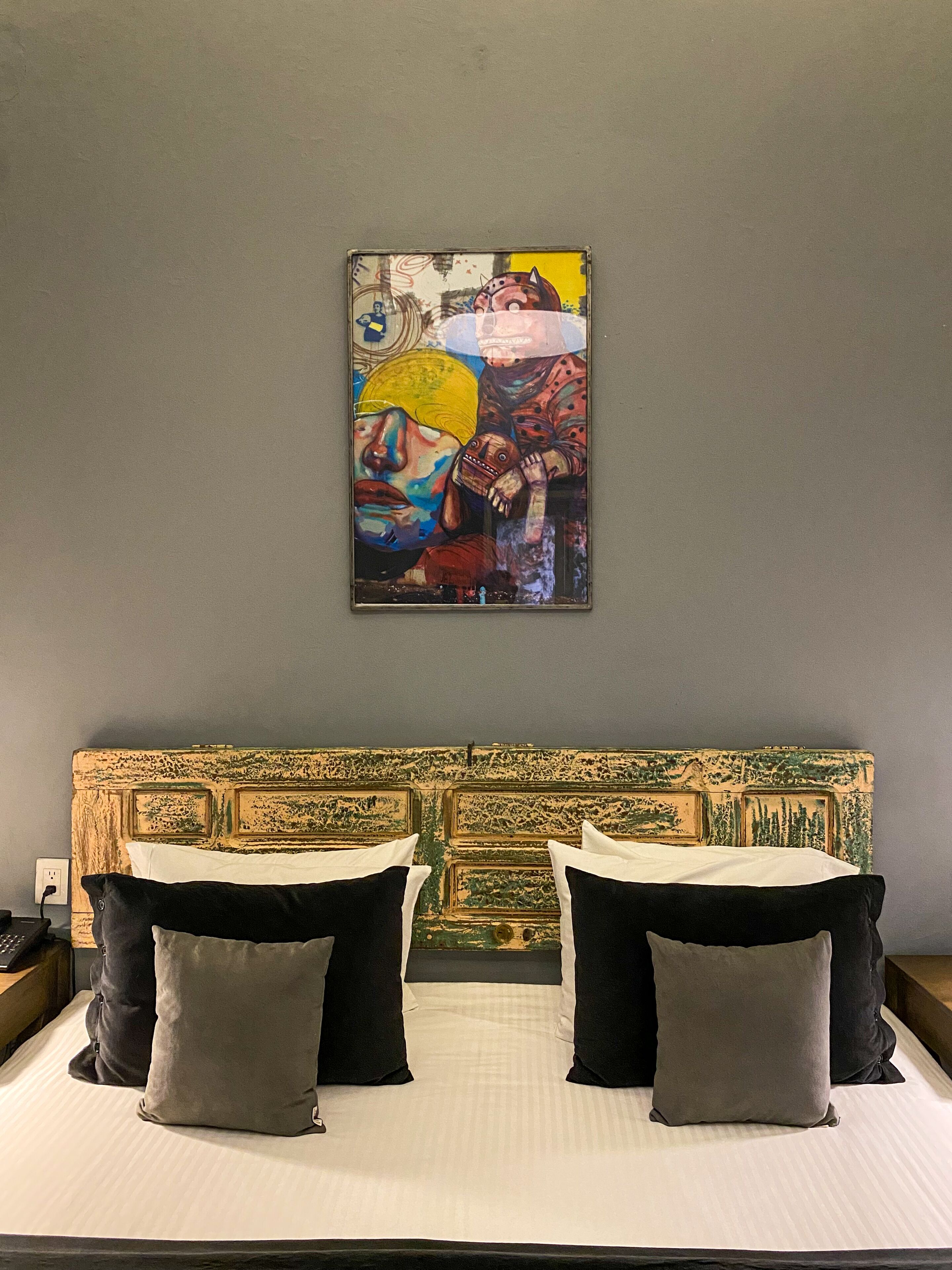 Habitación de hotel con una cama, dos almohadas, cabecera con diseño pintado y una obra de arte colorida en la pared.