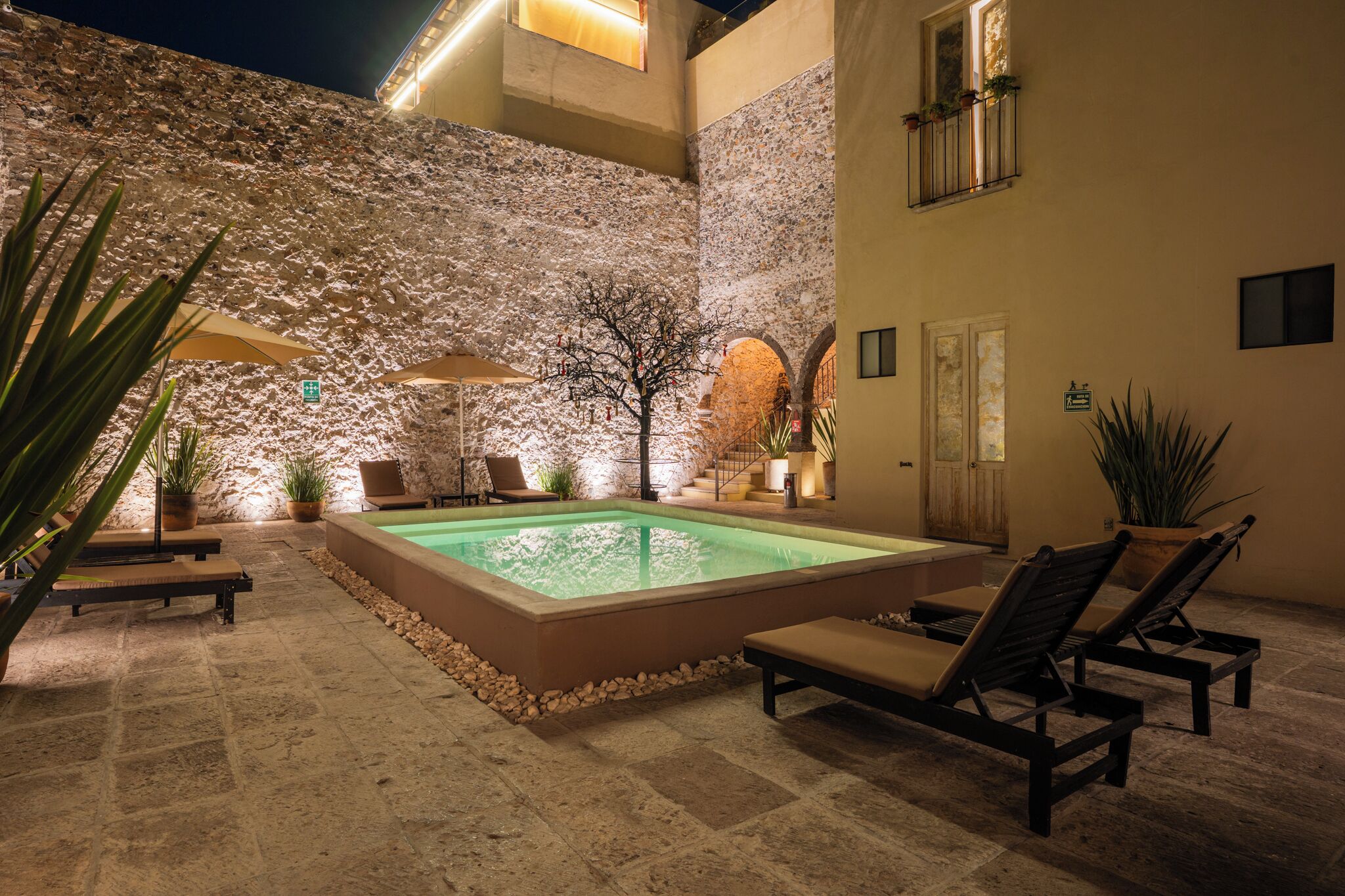 Zona de piscina al aire libre con paredes de piedra, sillas de descanso y sombrillas.