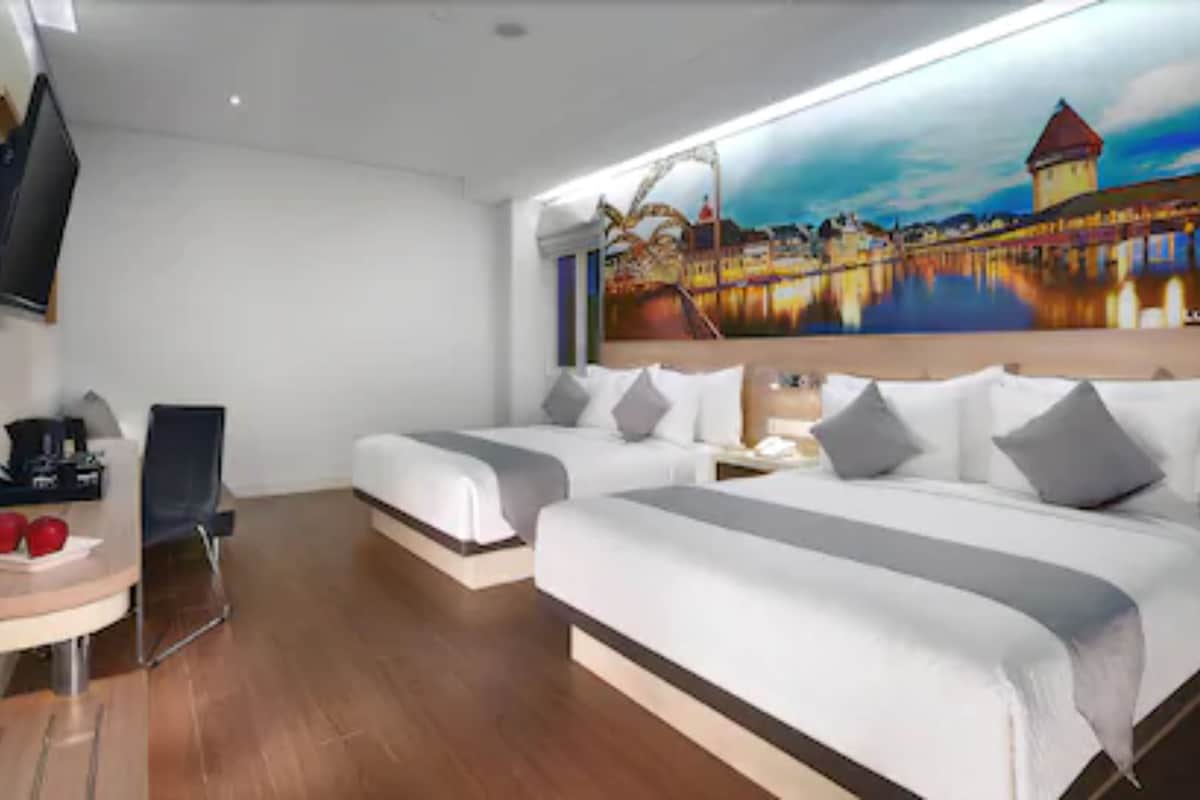 Une chambre d’hôtel avec deux lits, un bureau, une chaise, une télévision et un tableau représentant un paysage urbain.