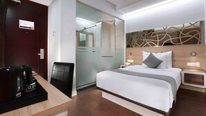 Ein Hotelzimmer mit Bett, Nachttischen, einem Schreibtisch mit Kaffeemaschine, einem Stuhl und einer Glastür zum Badezimmer.