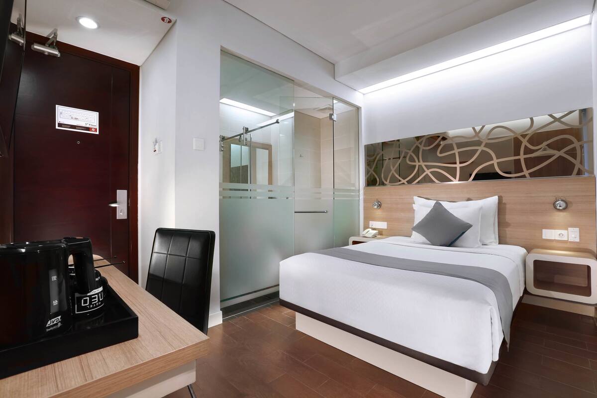 Habitación de hotel con cama, mesitas de noche, escritorio con cafetera, una silla y puerta de vidrio que da al baño.