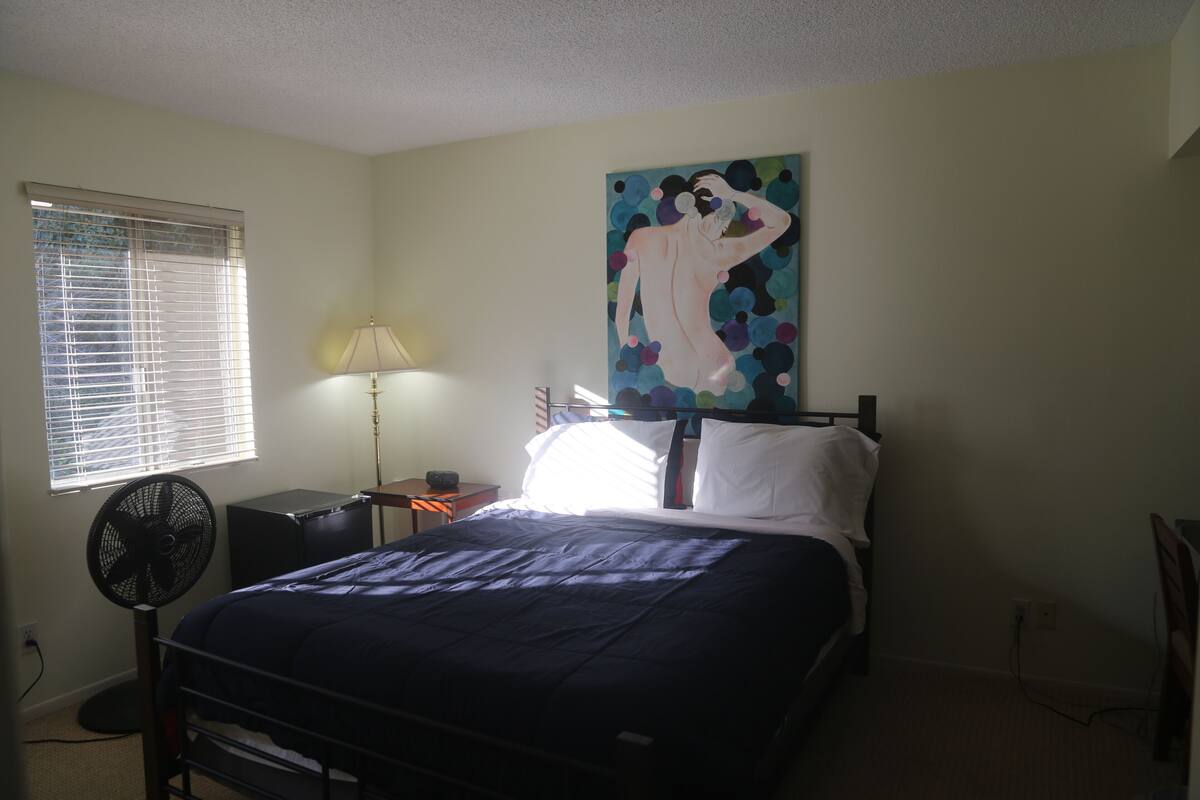 A bedroom with a bed, a standing fan, a lamp, a painting, and a window with blinds.