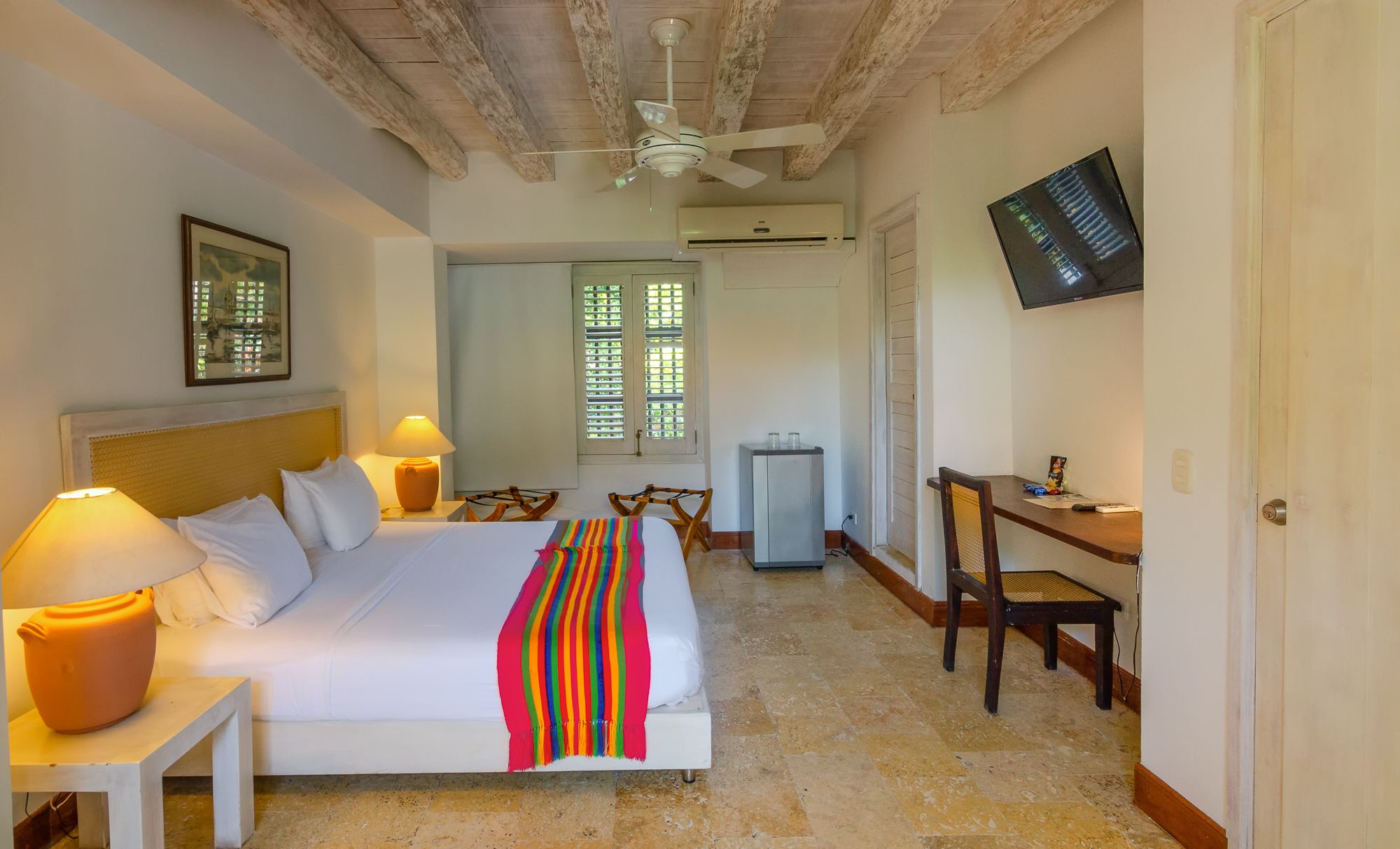 Una habitación de hotel con cama, lámparas de noche, escritorio, silla, televisor y aire acondicionado.