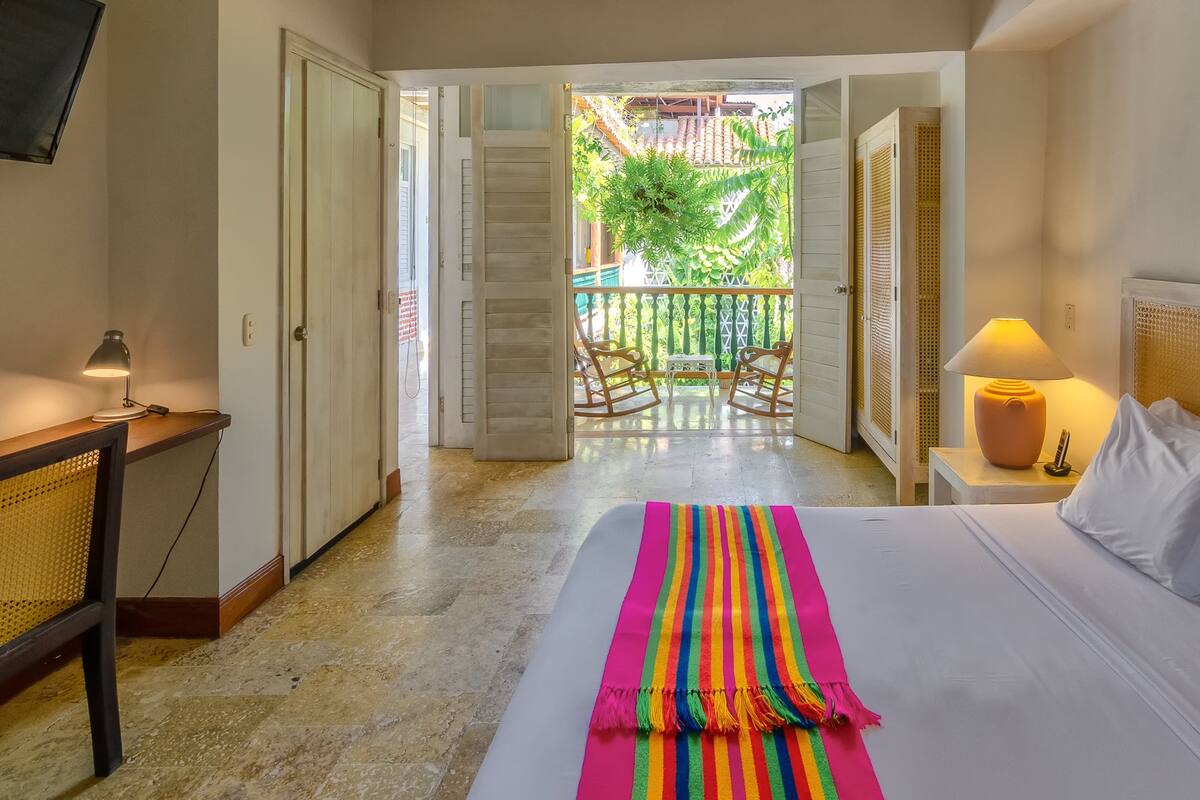 Habitación de hotel con cama, escritorio con lámpara, silla y vista a un balcón con barandilla y vegetación.