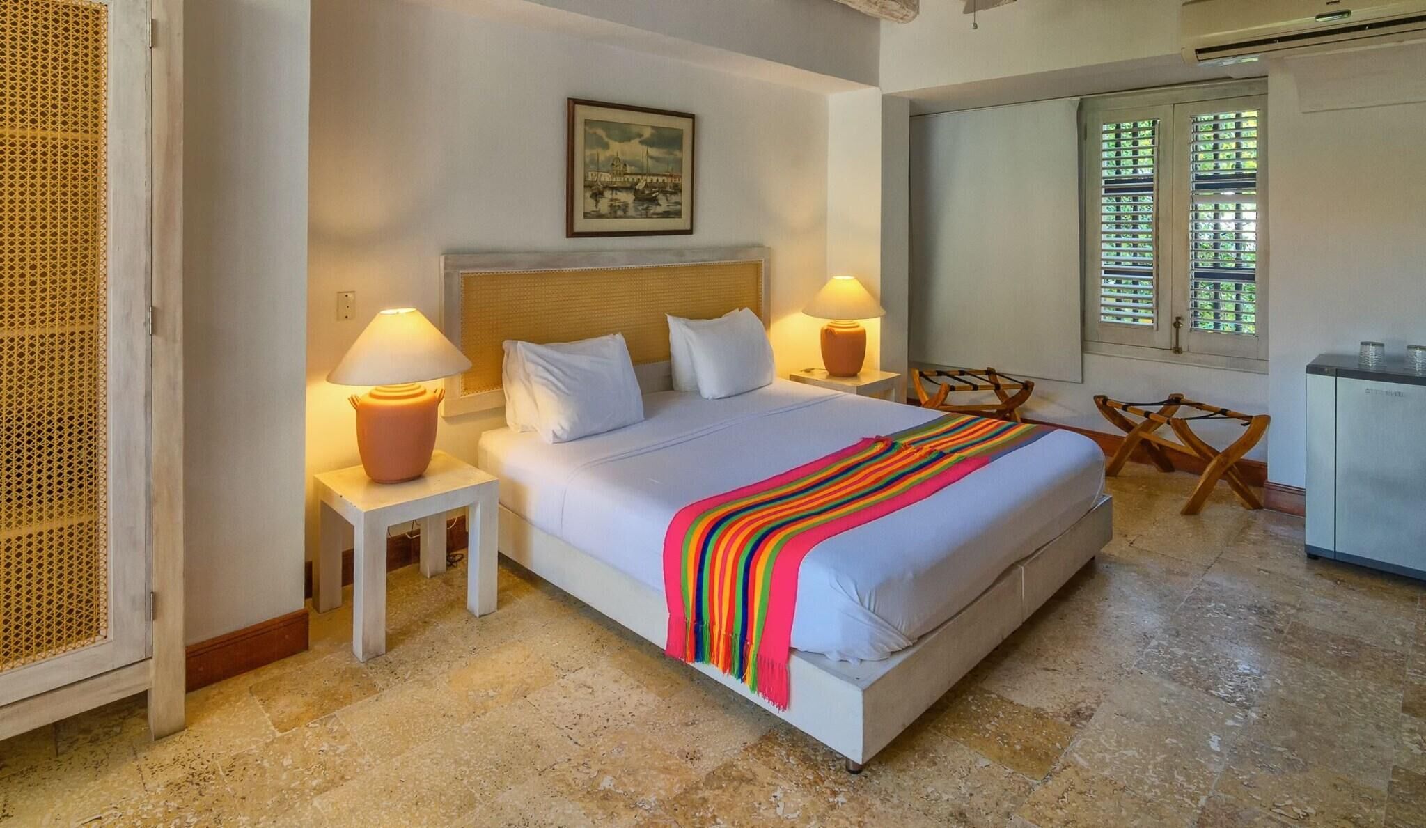 Une chambre d’hôtel avec un lit, deux lampes de chevet, un tableau au mur, une fenêtre avec des stores et une petite table avec une chaise.