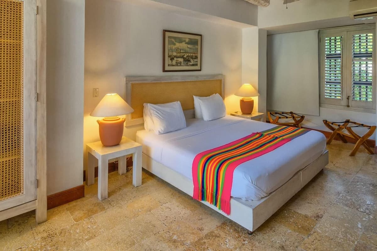Un cuarto de hotel con una cama, dos lámparas de noche, un cuadro en la pared, una ventana con persianas y una pequeña mesa con una silla.