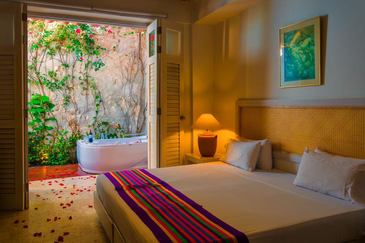 Habitación de hotel con cama, lámpara de noche, ventana grande con vistas a zonas verdes y bañera.