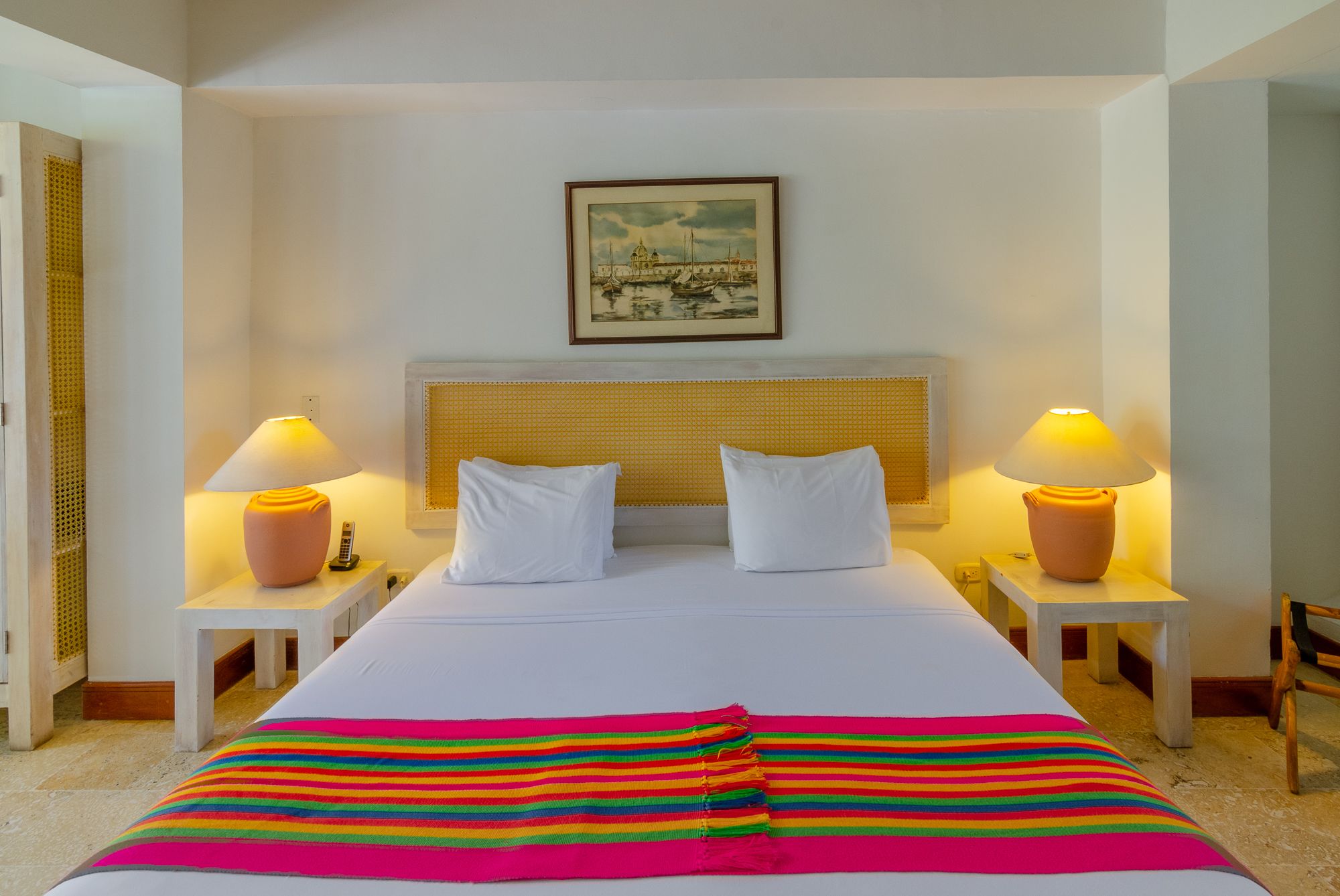 Habitación de hotel con una cama grande, dos lámparas de noche, un cabecero, un cuadro enmarcado en la pared y una manta colorida a rayas sobre la cama.