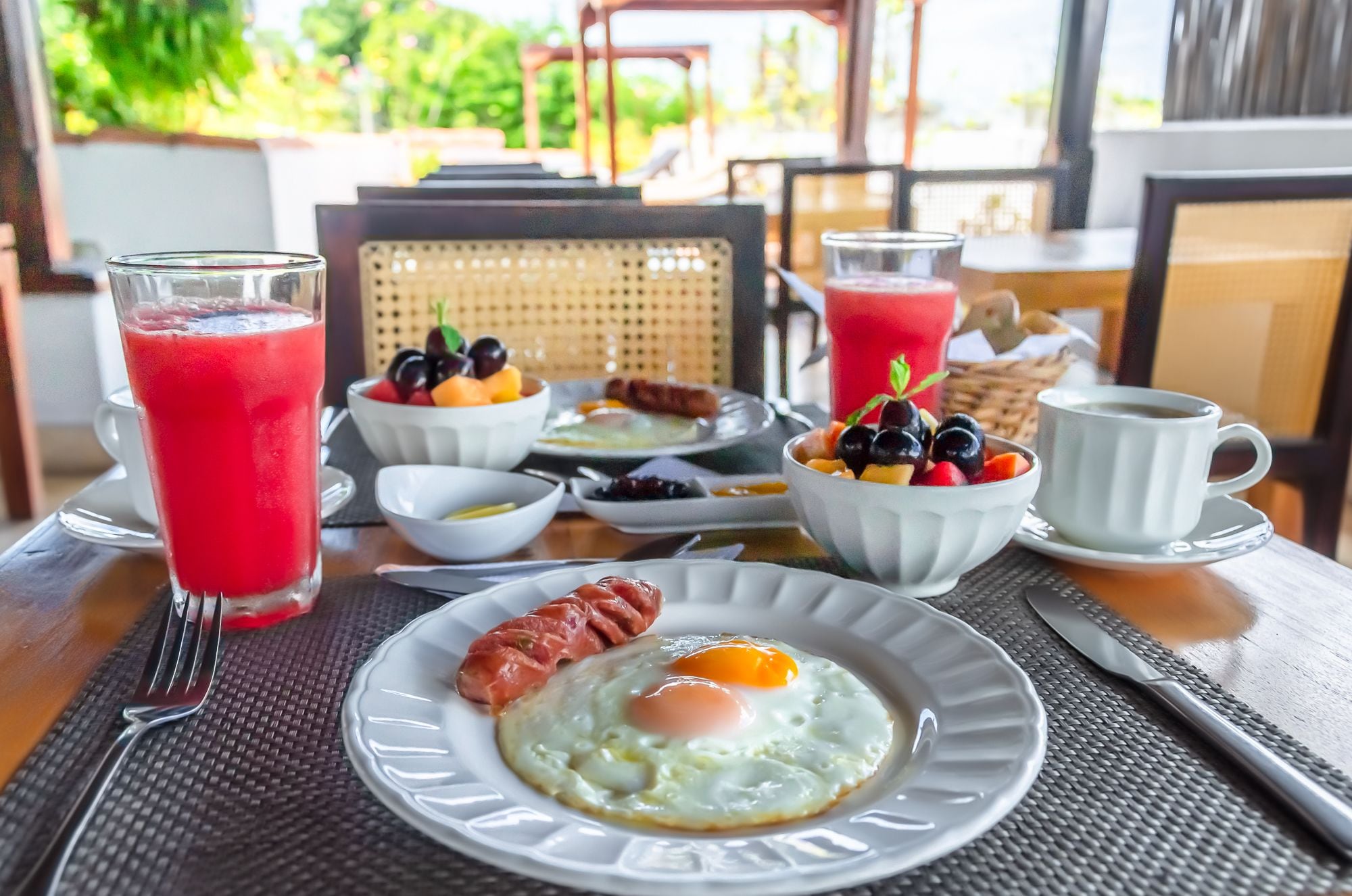 Une table dressée pour le petit-déjeuner, avec une assiette d’œufs, de saucisses et de fruits, ainsi que deux verres de jus rouge et une tasse de café.