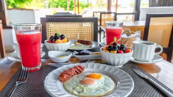 Une table dressée pour le petit-déjeuner, avec une assiette d’œufs, de saucisses et de fruits, ainsi que deux verres de jus rouge et une tasse de café.