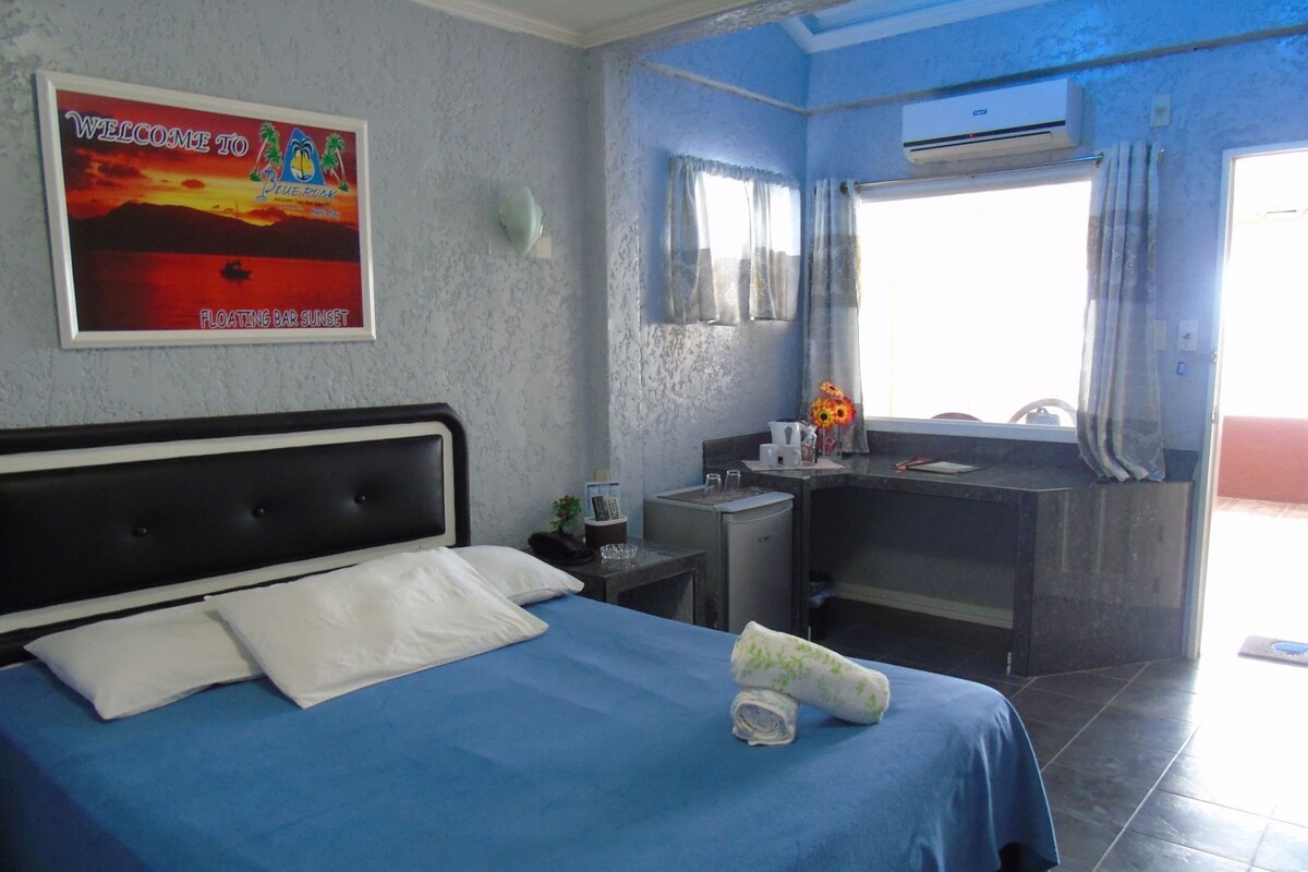 A hotel room with a bed, a desk, a small fridge, a window with curtains, and a poster on the wall.