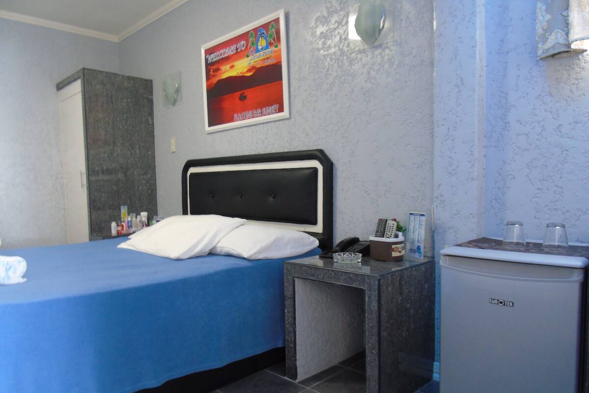 Habitación de hotel con cama, un pequeño escritorio con teléfono y controles remotos, un minibar y un cartel en la pared.