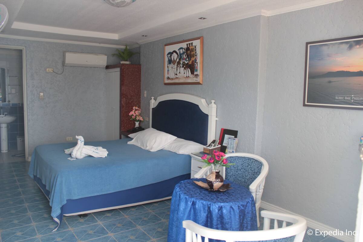 Una habitación de hotel con una cama, una mesita con un florero, una silla y un cuadro colgado en la pared.