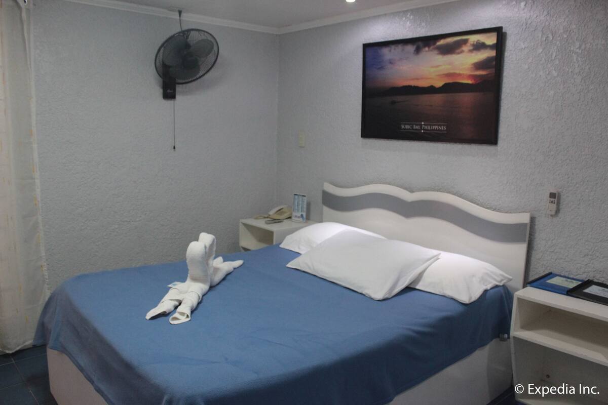 Habitación de hotel con cama, mesita de noche con teléfono, ventilador de pared y una imagen de una puesta de sol.