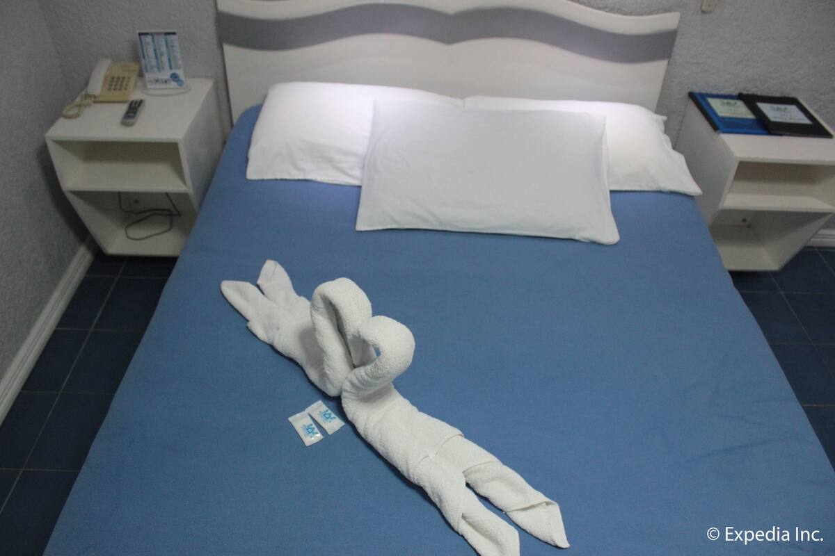 Habitación de hotel con cama, almohadas blancas, colcha azul, toalla y mesita de noche con teléfono y un libro.