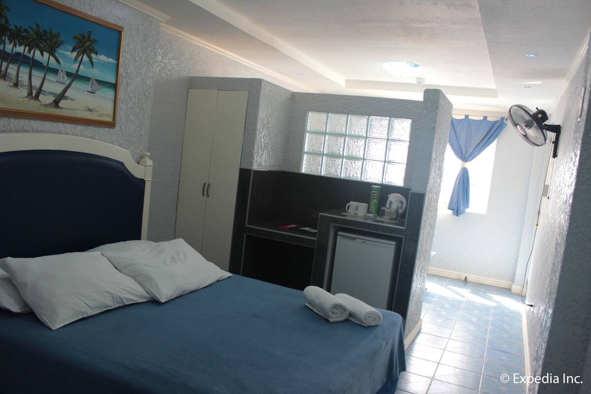Habitación de hotel con cama, escritorio, refrigerador pequeño, ventana con cortinas y un cuadro en la pared.