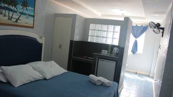 A hotel room with a bed, a desk, a small fridge, a window with curtains, and a painting on the wall.