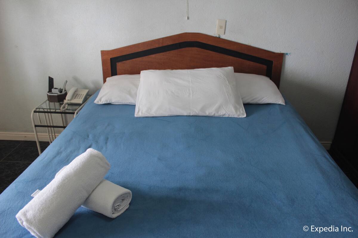 Una cama bien hecha con una colcha azul, dos almohadas blancas y una toalla blanca enrollada al lado.